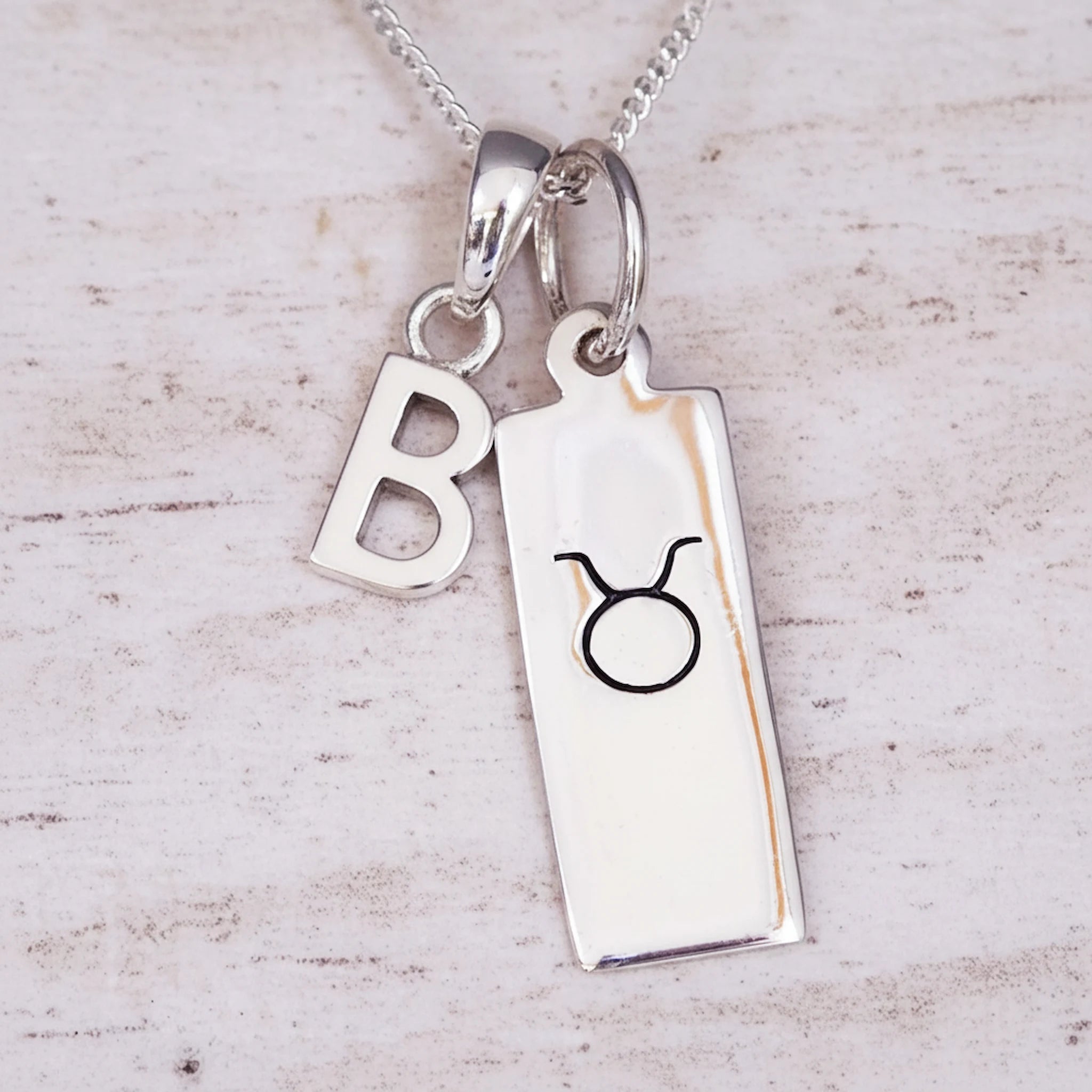 sterling Silver necklace with a 'B' initial pendant and a taurus zodiac star sign pendant on a light wooden background.