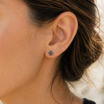 A close-up of a woman's ear wearing a small round stud earring with a dark gray center and silver-toned halo setting.