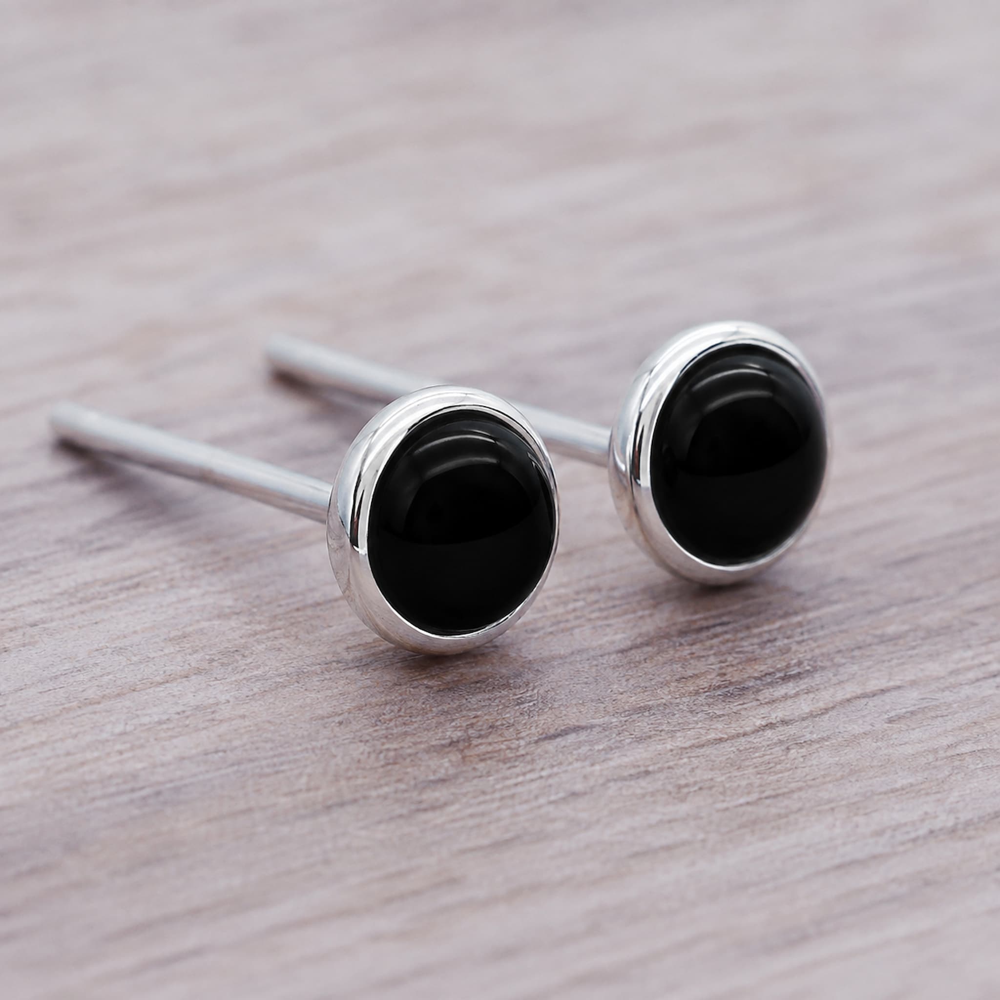 A pair of silver stud earrings with round black onyx cabochons, resting on a light brown wood grain surface.
