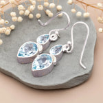 Silver earrings with teardrop-shaped blue gemstones on a stone surface with dried flowers.
