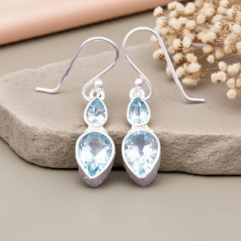 Silver earrings with blue teardrop-shaped gemstones on a stone surface with dried flowers.