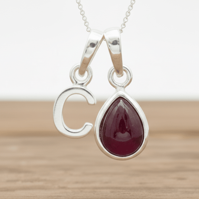 Silver c initial necklace with a teardrop-shaped red ruby gemstone pendant on a wooden surface.