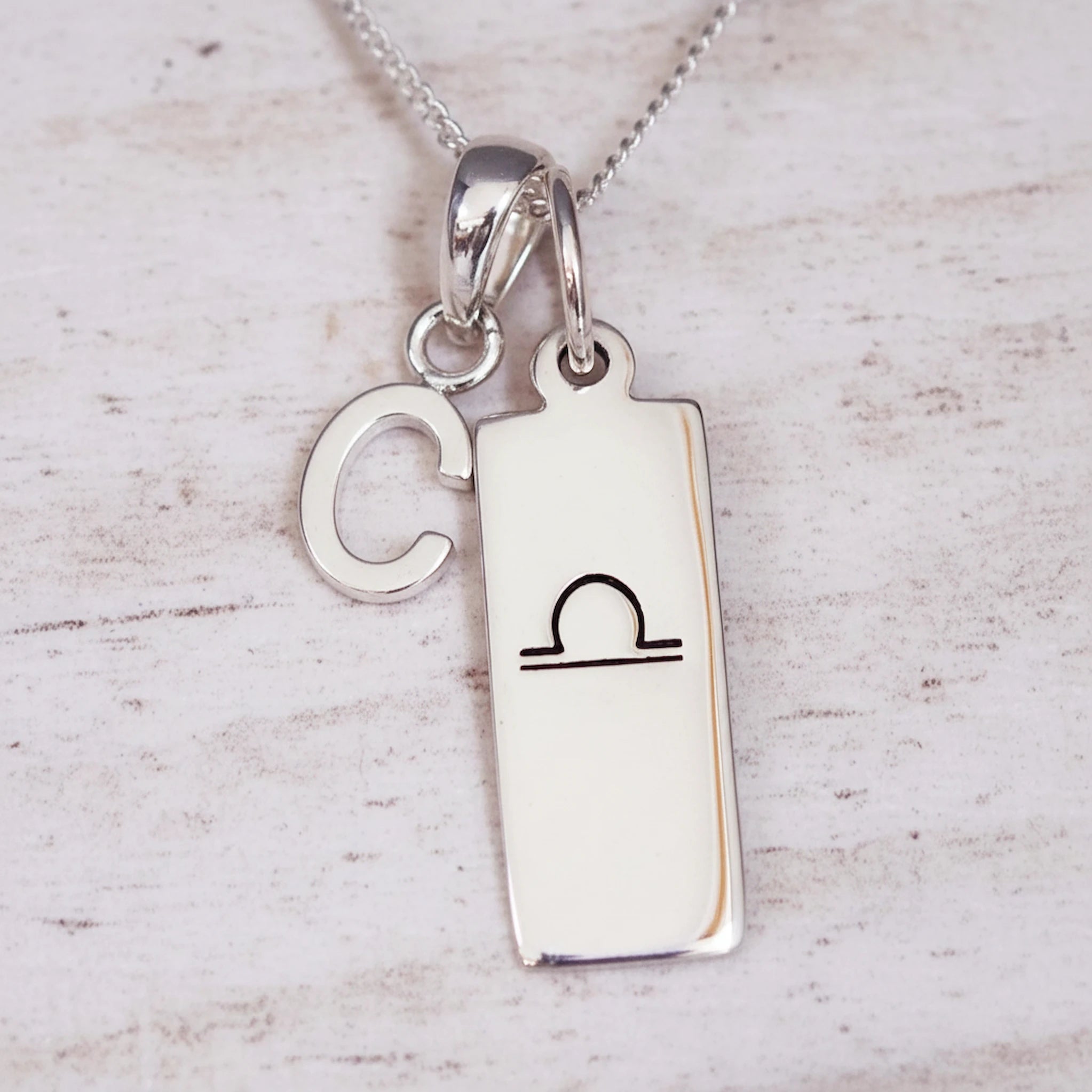 Silver necklace with a pendant featuring a letter 'C' and a libra zodiac symbol on a light wooden background.