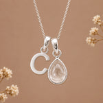 A silver initial pendant necklace featuring a polished letter 'C' and a teardrop-shaped clear gemstone pendant, displayed against a warm brown background with delicate dried flowers.
