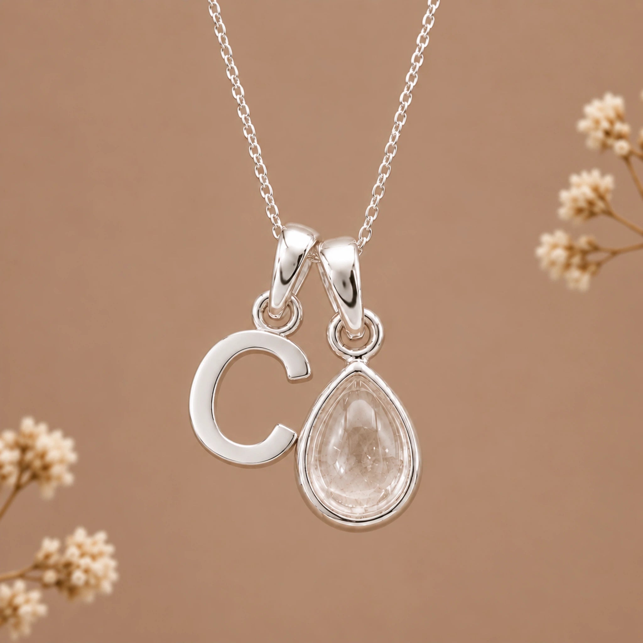 A silver initial pendant necklace featuring a polished letter 'C' and a teardrop-shaped clear gemstone pendant, displayed against a warm brown background with delicate dried flowers.