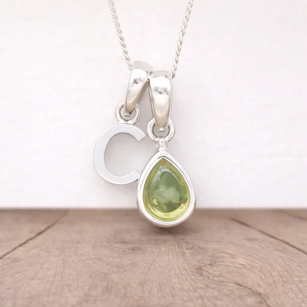 Silver necklace with a 'C' letter pendant and a green peridot teardrop pendant on a wooden surface