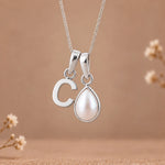 A silver initial necklace featuring a lowercase-style letter 'c' pendant and a teardrop-shaped white pearl pendant on a delicate silver chain, set against a warm beige background with subtle dried floral decorations.