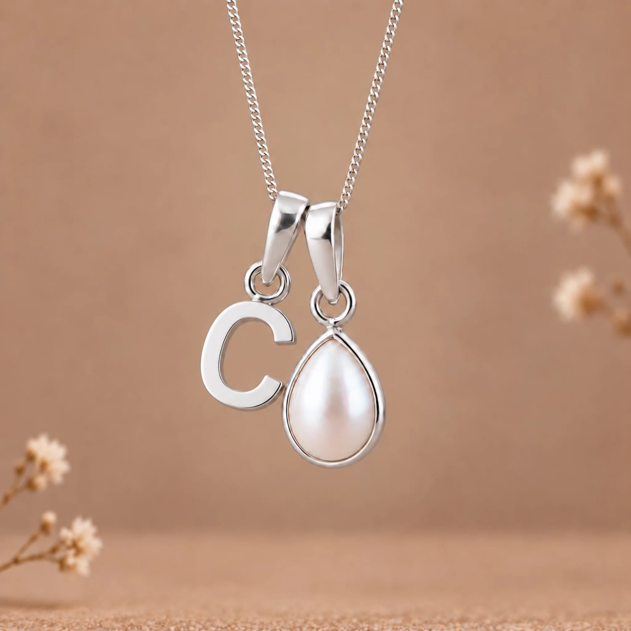 A silver initial necklace featuring a lowercase-style letter 'c' pendant and a teardrop-shaped white pearl pendant on a delicate silver chain, set against a warm beige background with subtle dried floral decorations.