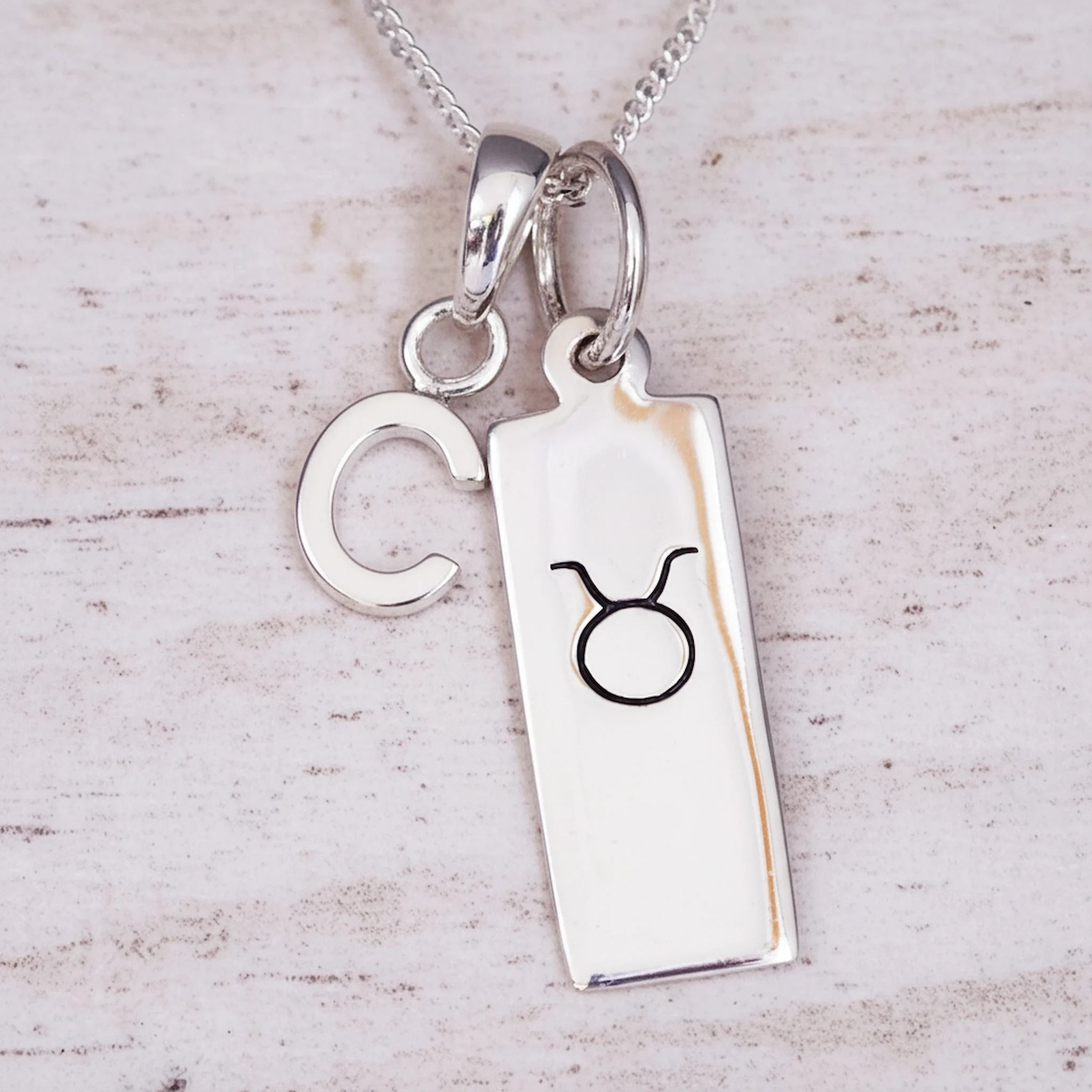 sterling Silver necklace with a 'C' initial pendant and a taurus zodiac star sign pendant on a light wooden background.