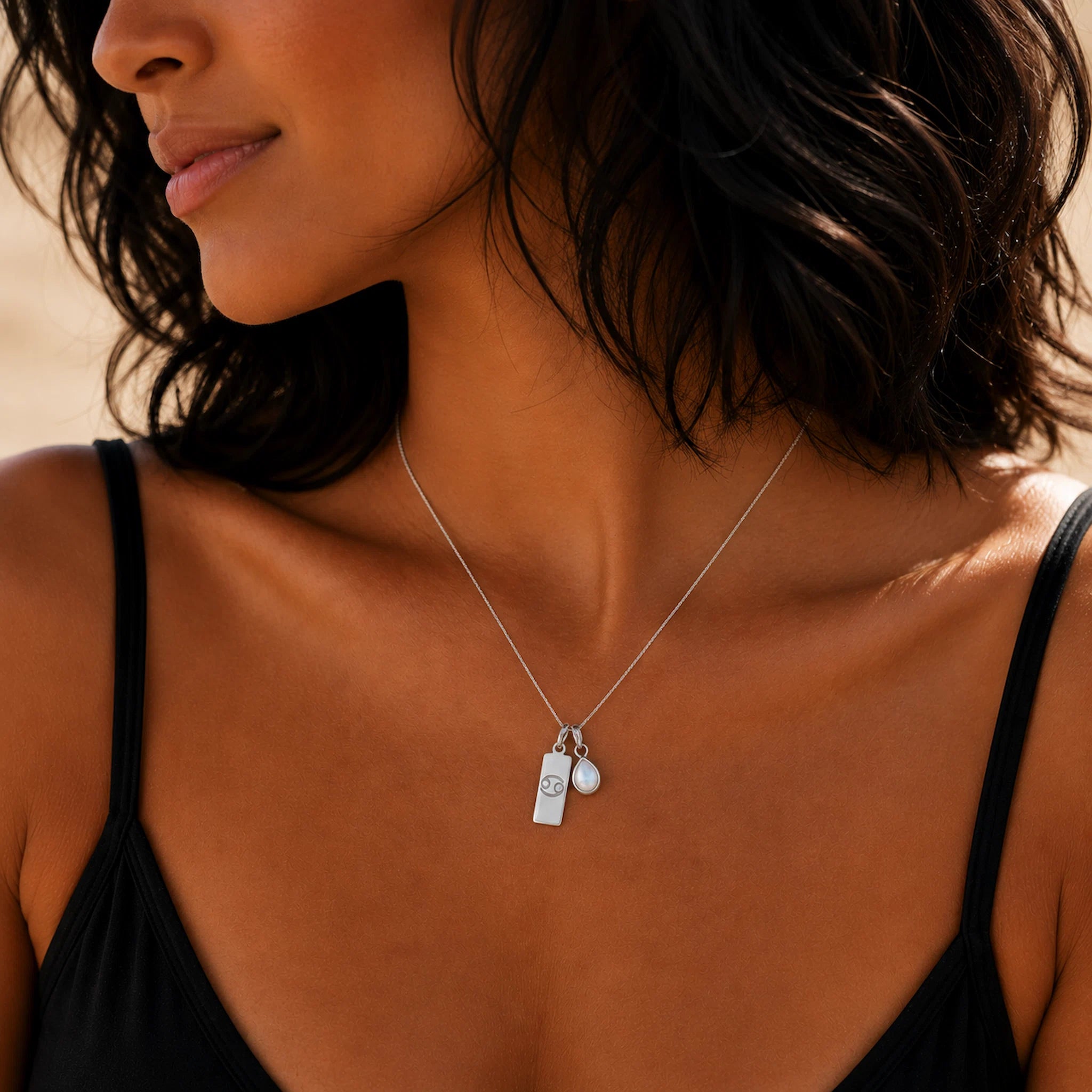 Close-up of a woman wearing a silver necklace with a tear drop june birthstone pearl pendant and a cancer zodiac line glyph pendant.