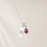 sterling silver necklace with a teardrop july birthstone ruby pendant and cancer line glyph pendant hanging on a light background.
