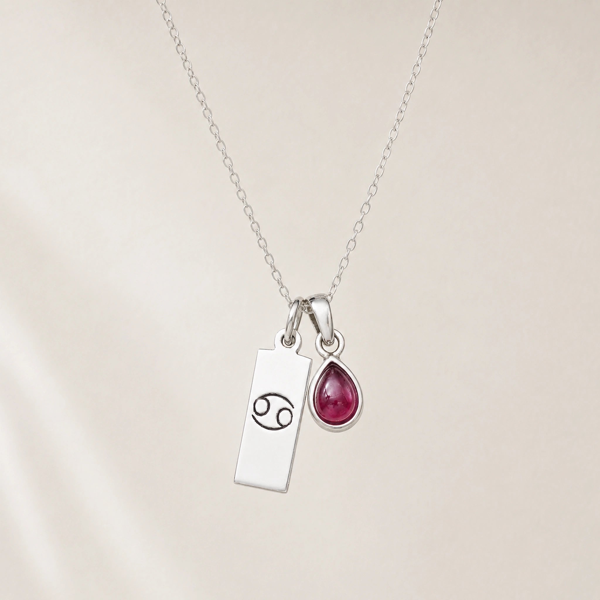 sterling silver necklace with a teardrop july birthstone ruby pendant and cancer line glyph pendant hanging on a light background.