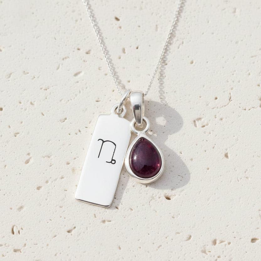 Silver necklace with a teardrop-shaped garnet gemstone pendant and a rectangular tag with a Capricorn zodiac symbol on a white background.