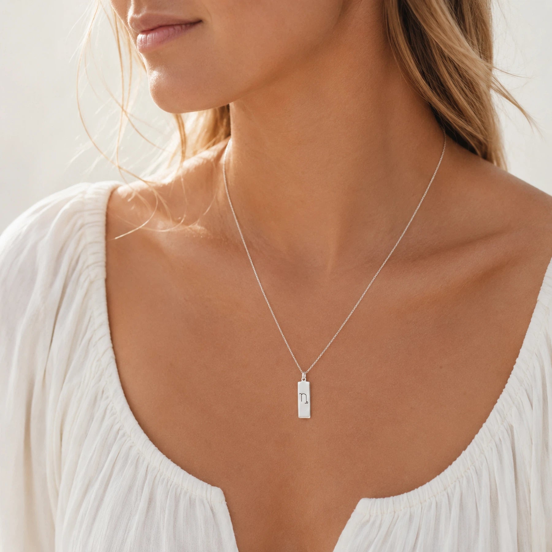 model wearing a sterling silver fine chain necklace with a rectangle pendant engraved with the capricorn zodiac line glyph symbol.