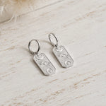 silver hoop earrings with rectangle charms with celestial design on a rustic wooden surface