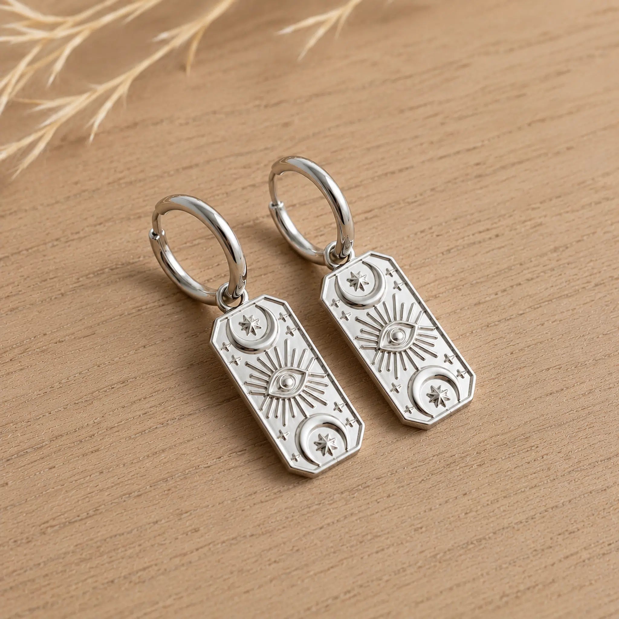 A pair of silver-toned rectangular drop earrings with engraved celestial motifs, including suns, moons, and an eye of providence, resting on a light wood surface.