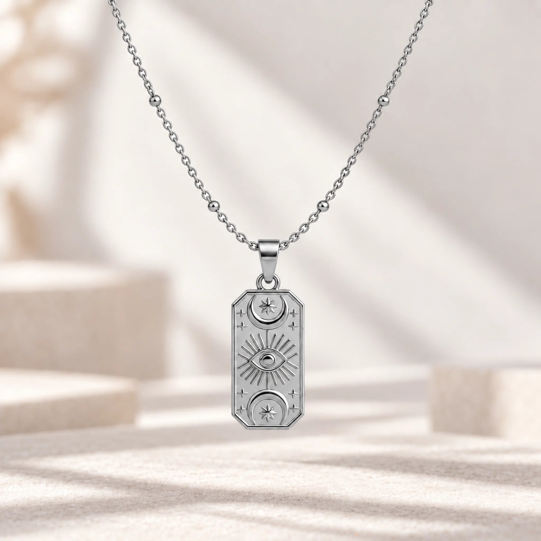 A silver-toned necklace with a rectangular pendant featuring celestial motifs including an eye of providence, sun rays, and crescent moons, displayed on a soft beige surface with soft shadows.