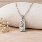 A silver-toned rectangular pendant necklace with embossed celestial motifs, resting on a light beige stone against a light wood surface with small dried flowers at the side.