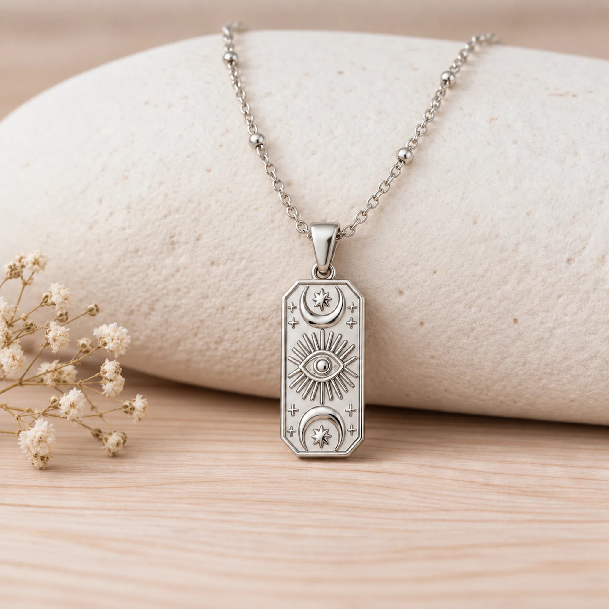 A silver-toned rectangular pendant necklace with embossed celestial motifs, resting on a light beige stone against a light wood surface with small dried flowers at the side.