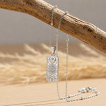 A silver engraved pendant necklace with celestial symbols hangs from a silver chain draped over a rustic wooden branch on a warm beige surface.