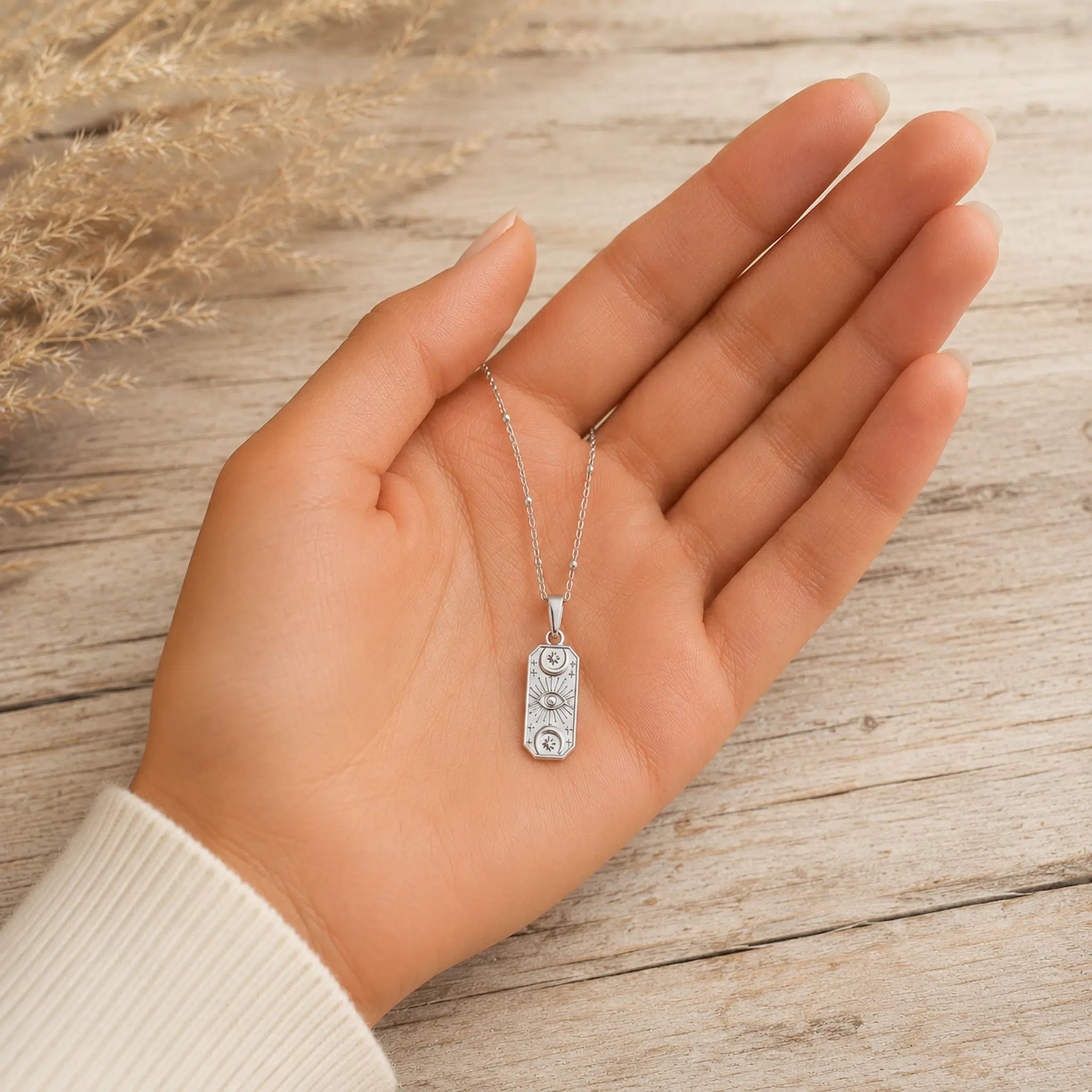 A silver-toned rectangular pendant necklace with circular cutout details, displayed on an open hand over a light wood surface with dried grass in the background.