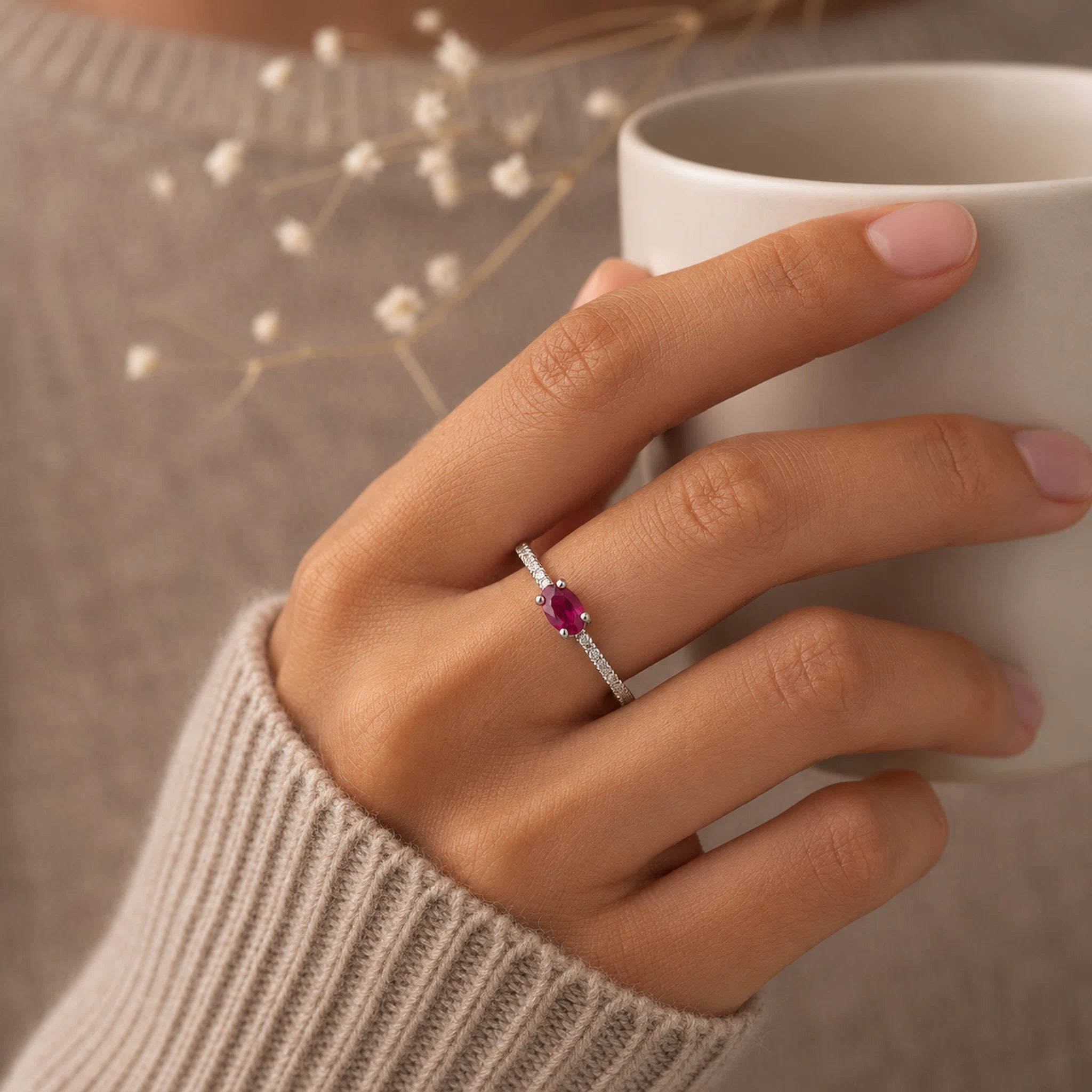 Hand holding a white mug with a sterling silver cubic zirconia pave stacking ring featuring a red ruby gemstone, wearing a beige sweater.