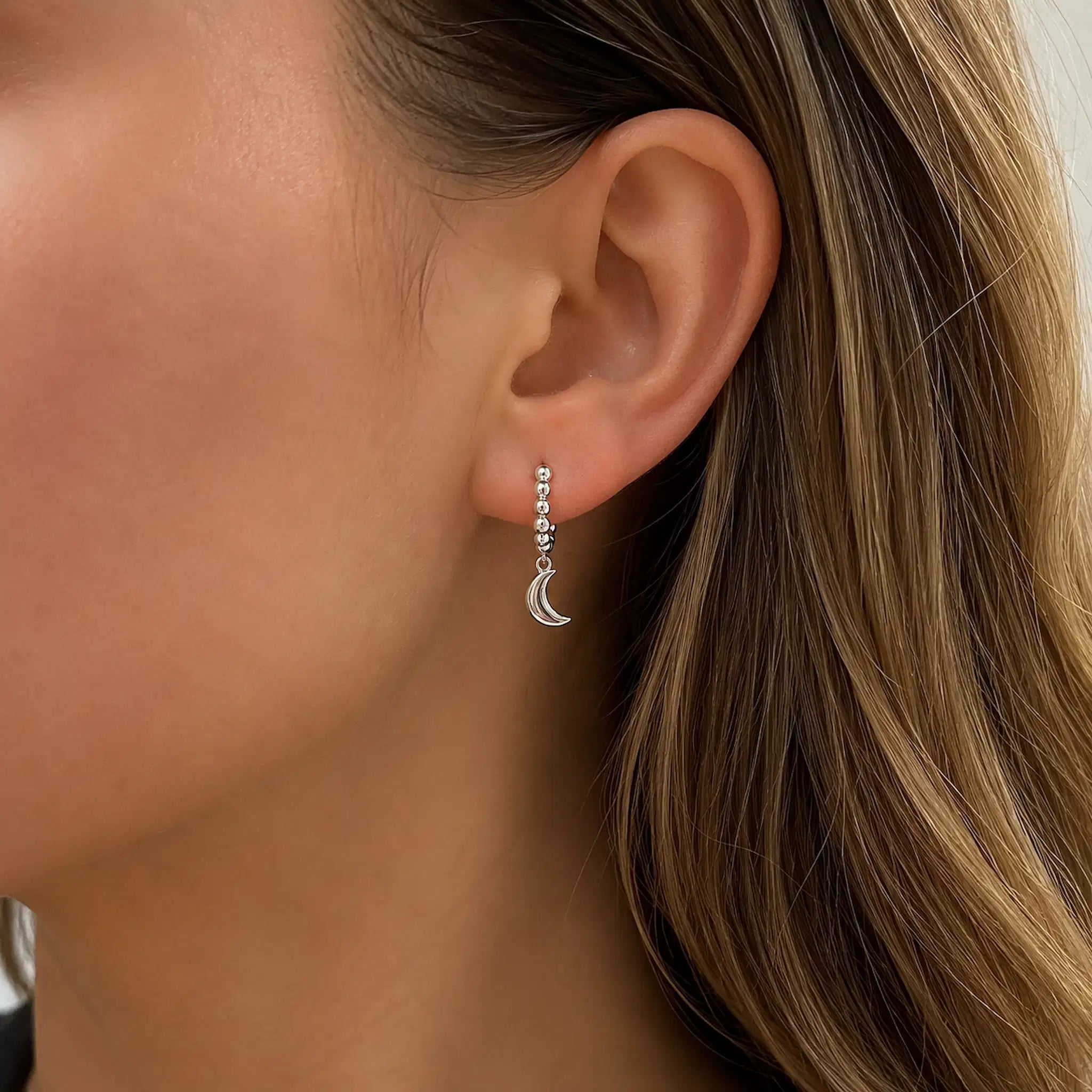 A close-up of a woman's ear wearing a delicate silver dangle earring with a small dangling crescent moon charm.