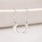 A pair of silver crescent moon dangle earrings with beaded hoops, hanging from a cream-colored display stand against a soft beige background.
