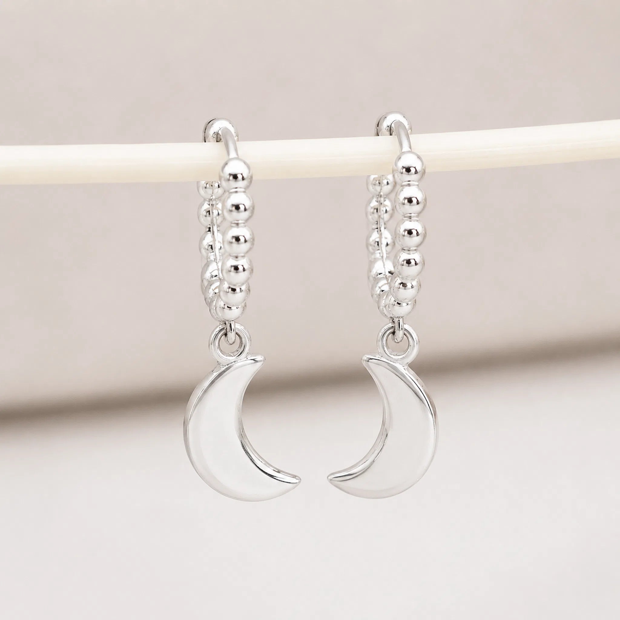 A pair of silver crescent moon dangle earrings with beaded hoops, hanging from a cream-colored display stand against a soft beige background.