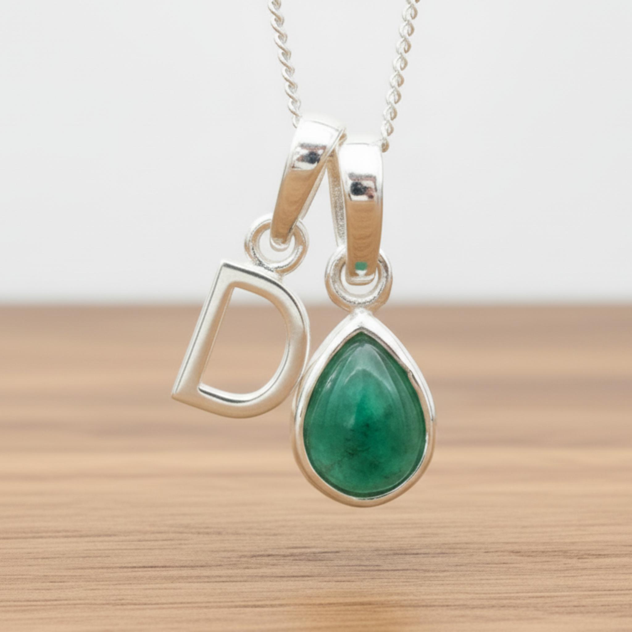 Silver Initial D necklace with a teardrop-shaped Emerald gemstone pendant hanging above a rustic wooden surface