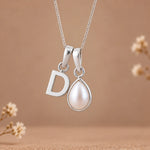 A silver necklace with a letter 'D' initial pendant and a teardrop-shaped pearl pendant, displayed against a warm brown background with subtle dried floral decorations.