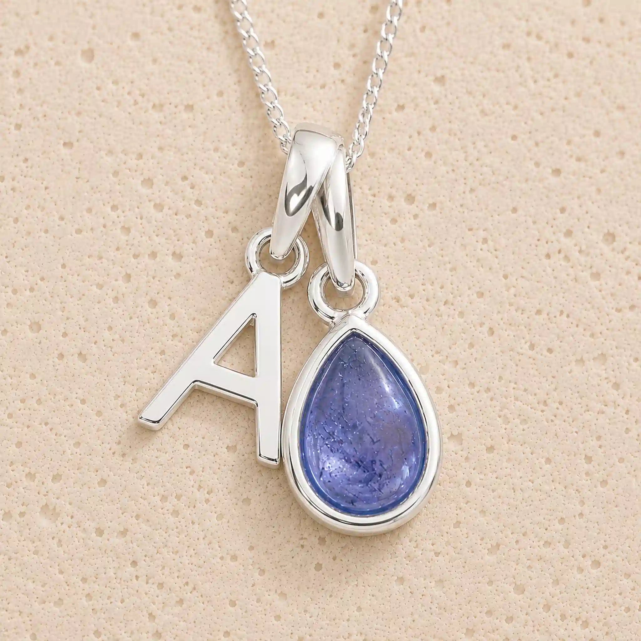Sterling Silver december birthstone necklace with a teardrop-shaped blue tanzanite gemstone pendant and letter 'A' charm on a beige background.