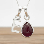 Silver necklace with a teardrop-shaped red ruby gemstone pendant and letter 'E' charm on a wooden surface.