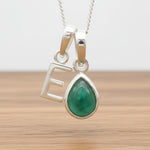 Silver Initial E necklace with a teardrop-shaped Emerald gemstone pendant hanging above a rustic wooden surface