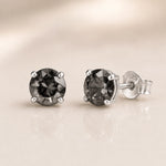 A pair of round-cut dark gray gemstone stud earrings in silver four-prong settings, with one earring facing forward and the other angled to show the silver post and butterfly back, set against a soft beige background with a subtle reflection.