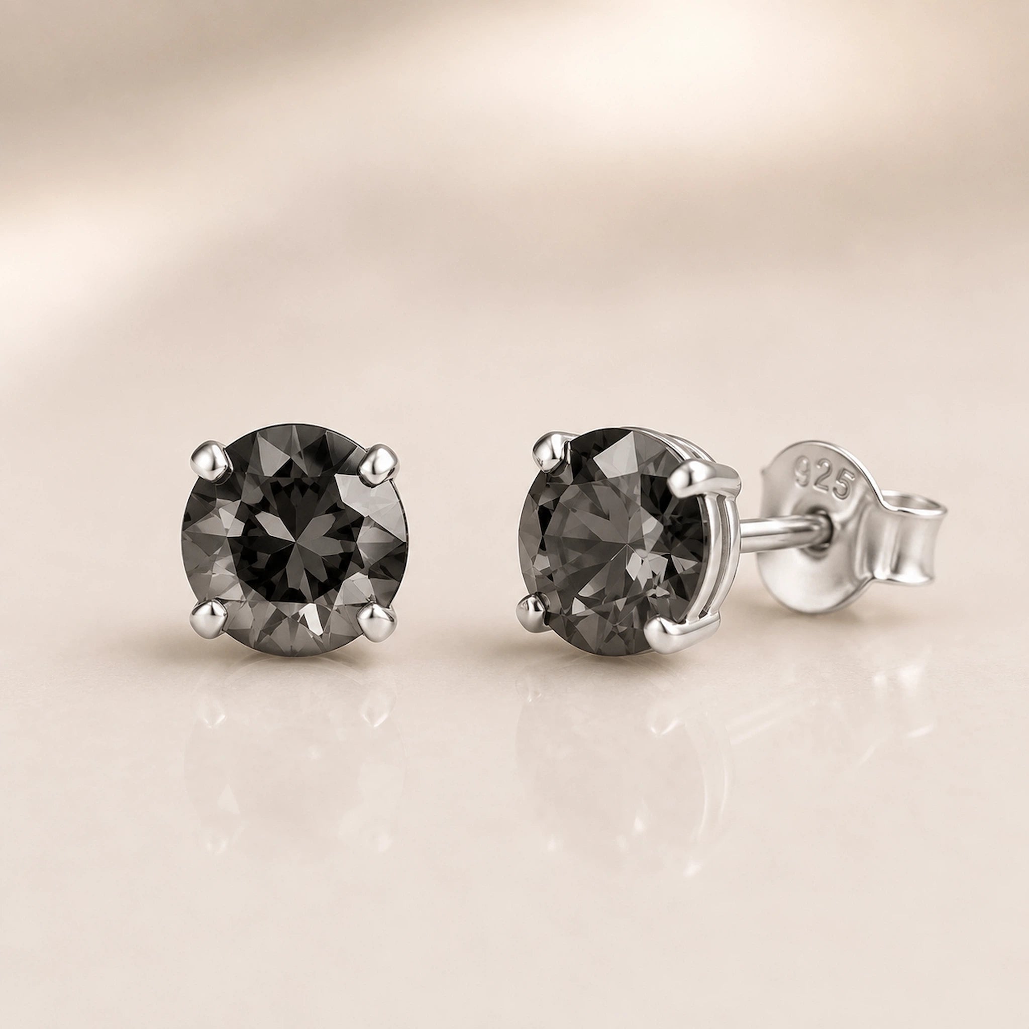 A pair of round-cut dark gray gemstone stud earrings in silver four-prong settings, with one earring facing forward and the other angled to show the silver post and butterfly back, set against a soft beige background with a subtle reflection.