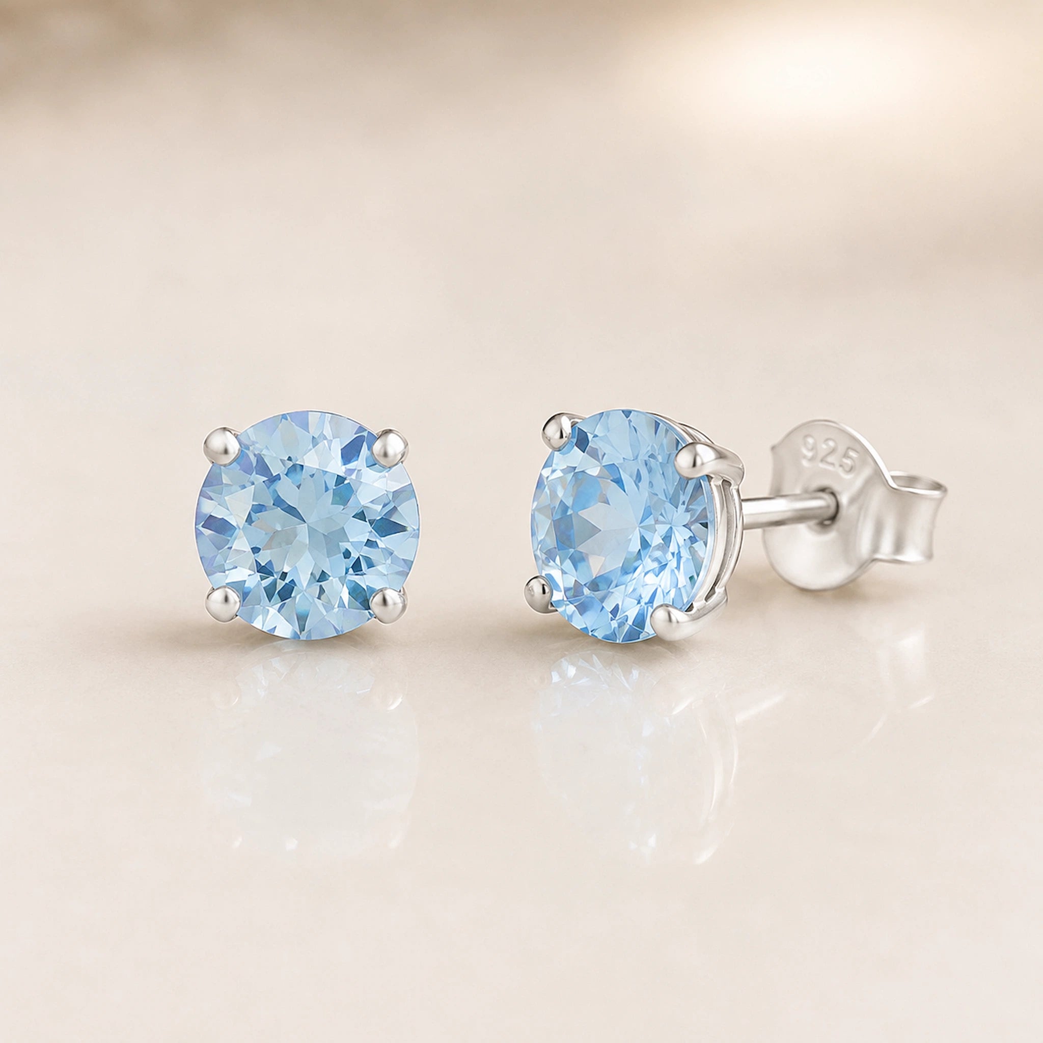 A pair of round-cut light blue gemstone stud earrings in a silver four-prong setting, with one earring facing forward and the other angled to show the post and backing engraved with '925'.