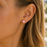 A close-up of a woman's ear wearing a small round stud earring with a pale pink stone.
