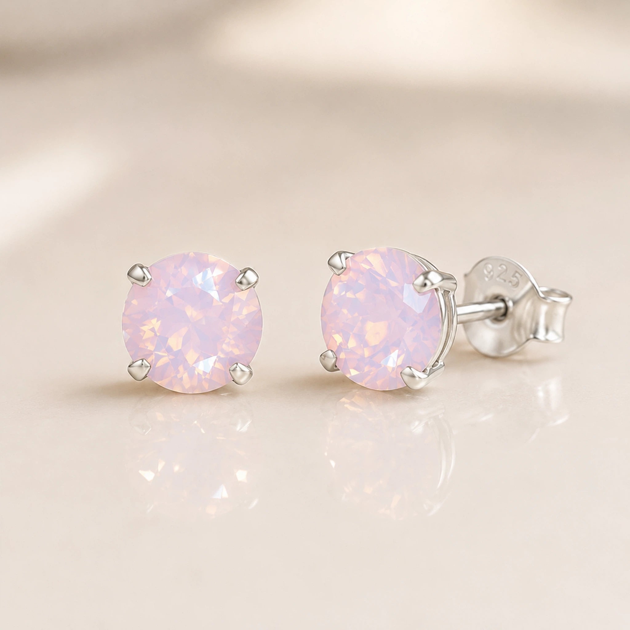 A pair of round-cut light pink opal stud earrings in silver prong settings, displayed on a soft beige reflective surface.