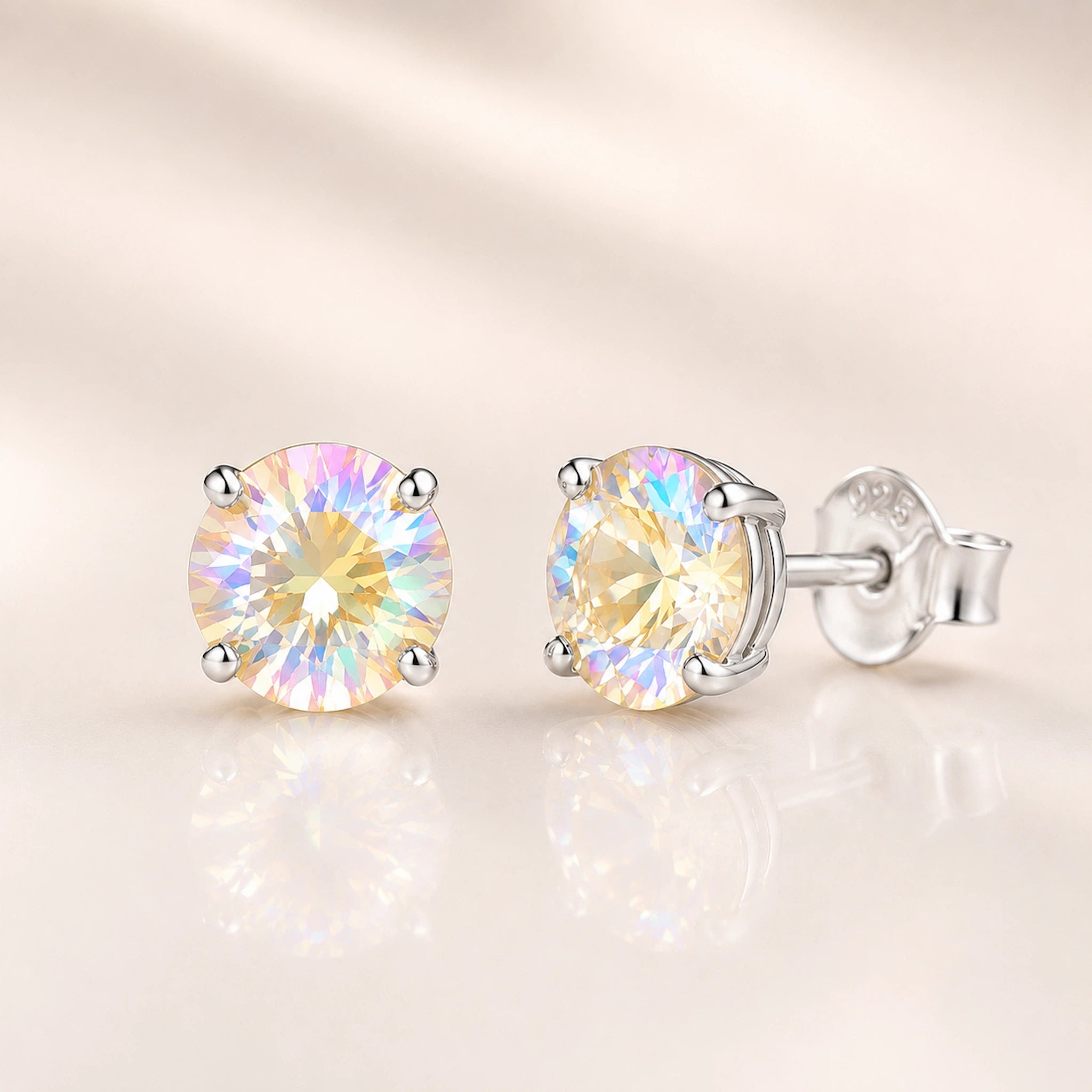 A pair of round-cut iridescent crystal stud earrings in silver four-prong settings, shown on a soft beige reflective surface.