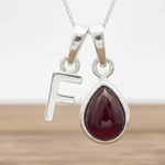 Silver necklace with a teardrop-shaped red ruby gemstone pendant and a silver 'F' charm on a wooden surface.