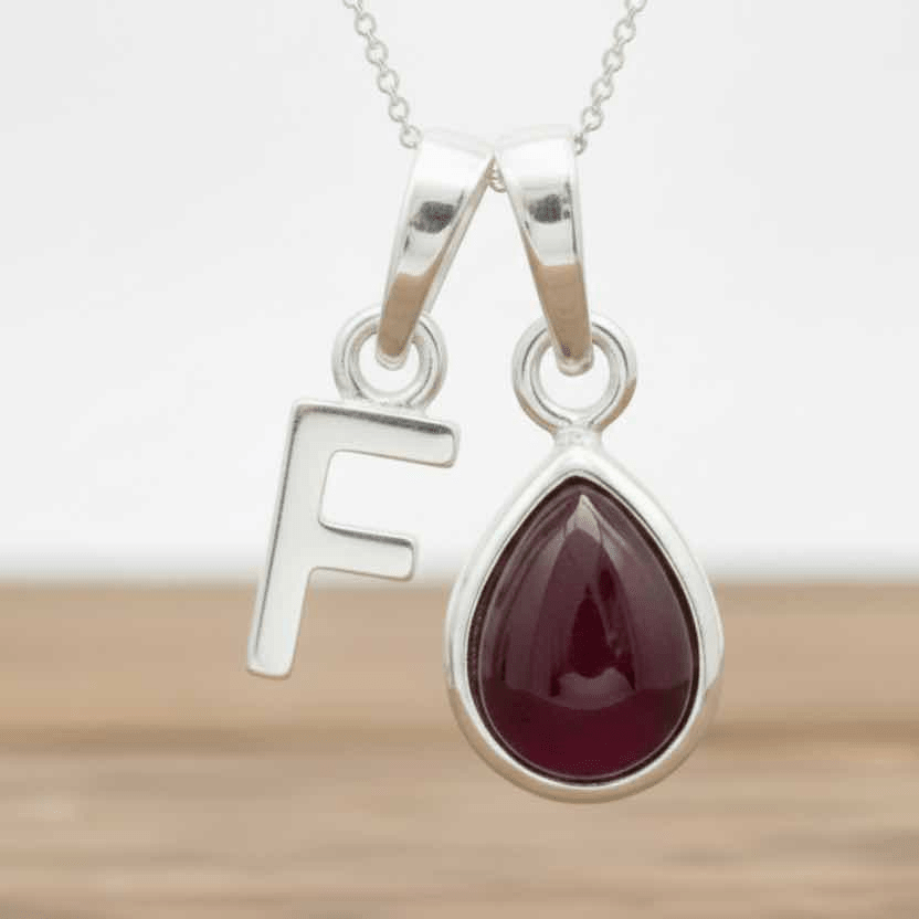 Silver necklace with a teardrop-shaped red ruby gemstone pendant and a silver 'F' charm on a wooden surface.
