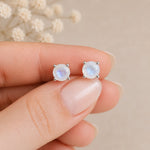 A close-up of a hand holding a pair of round silver stud moonstone earrings with iridescent natural faceted moonstones beze-set, set against a soft beige background with blurred dried flowers.