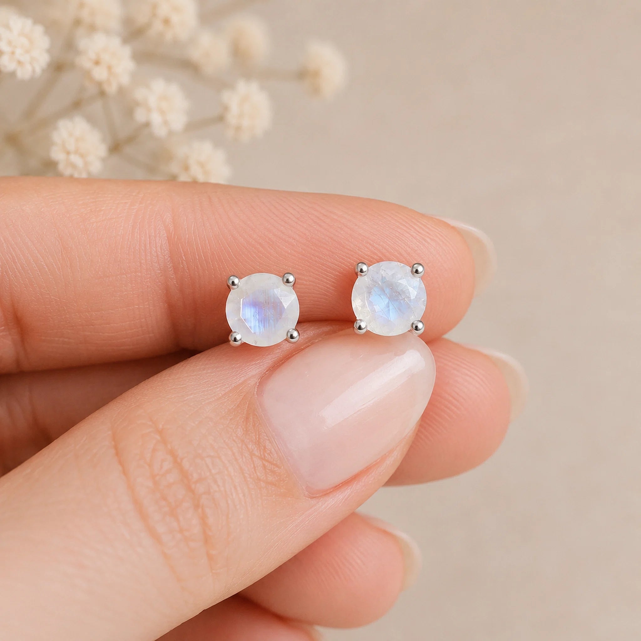 A close-up of a hand holding a pair of round silver stud moonstone earrings with iridescent natural faceted moonstones beze-set, set against a soft beige background with blurred dried flowers.