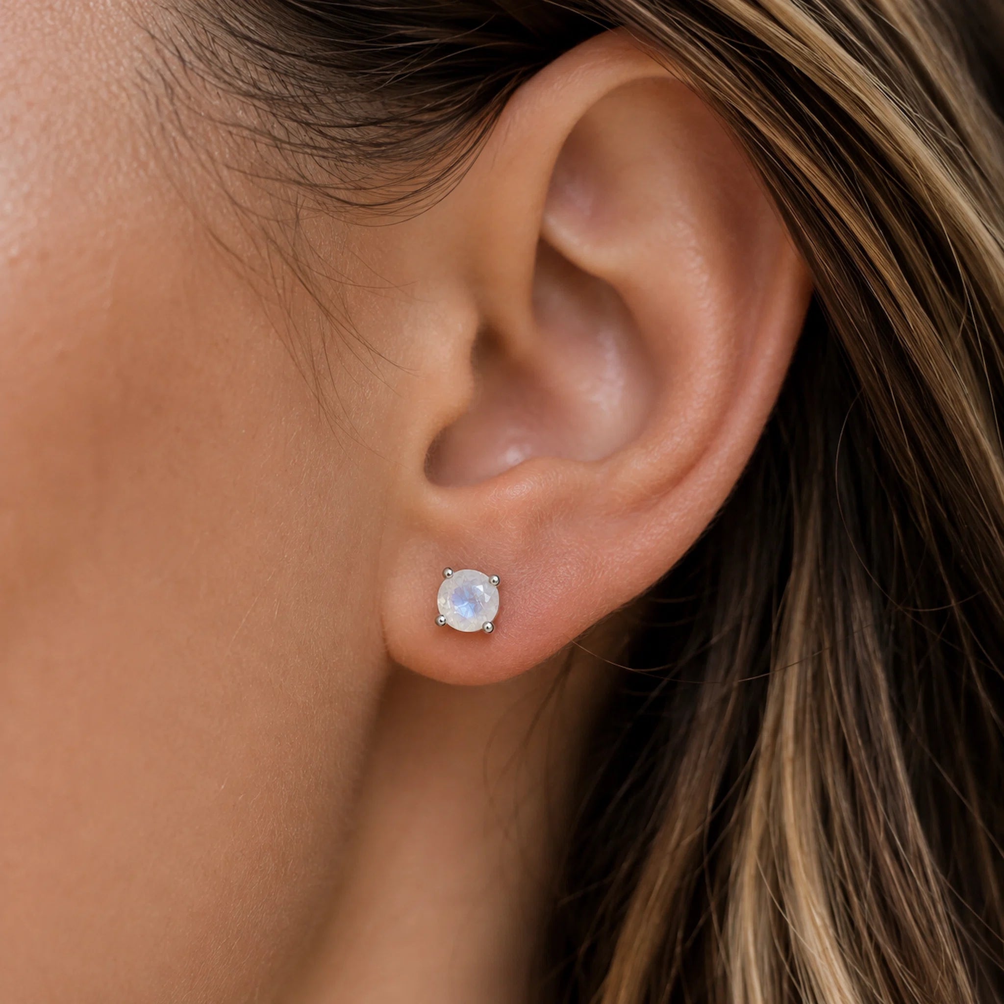 A close-up of a person's ear wearing a small stud moonstone earring with a round iridescent natural moonstone gemstone in a claw set silver-toned setting.