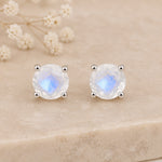 A pair of sterling silver faceted natural moonstone stud earrings with silver prongs, displayed on a beige stone surface with dried white flowers in the background.