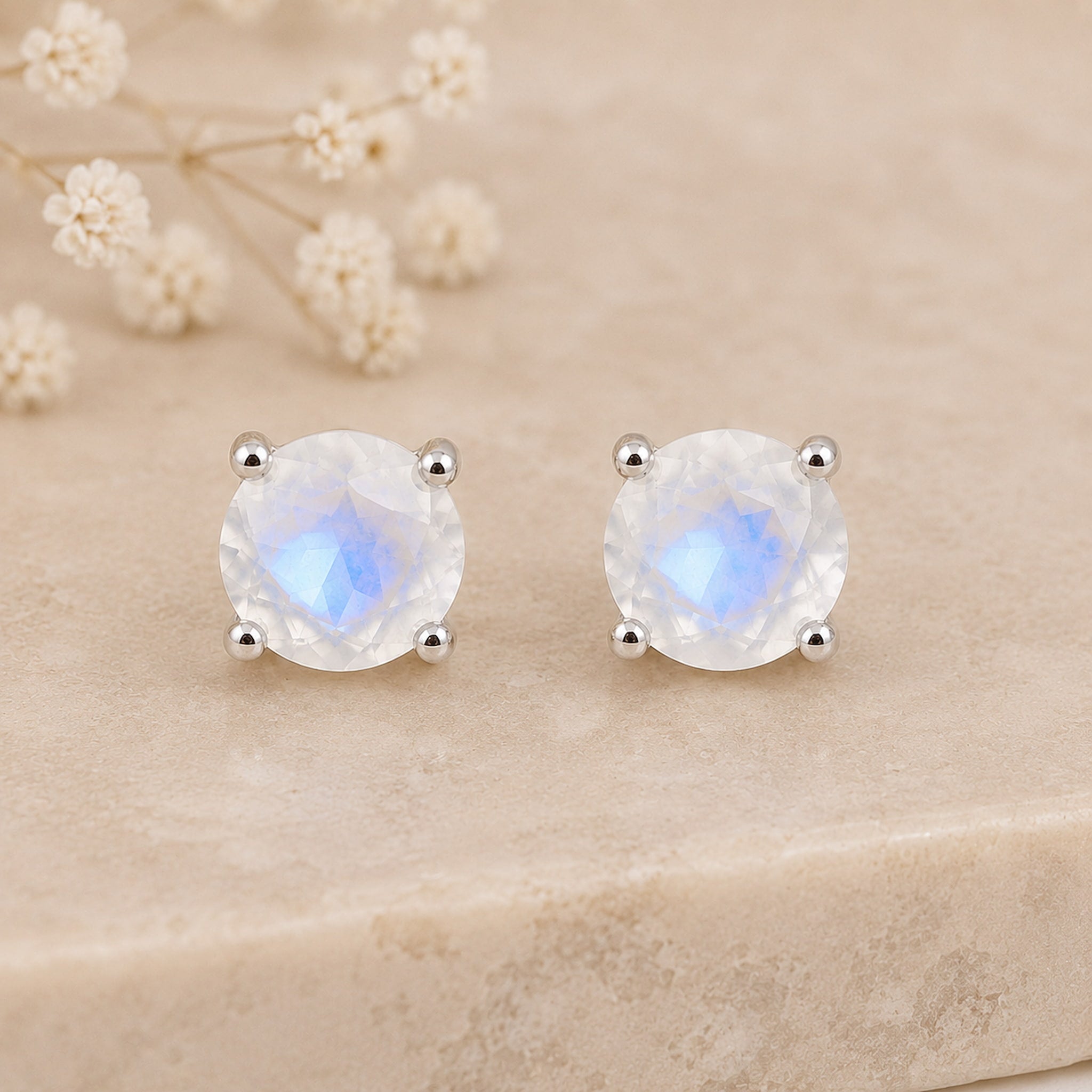 A pair of sterling silver faceted natural moonstone stud earrings with silver prongs, displayed on a beige stone surface with dried white flowers in the background.