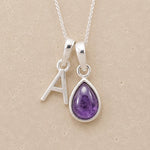 Silver necklace with a teardrop-shaped february birthstone amethyst gemstone pendant and a silver 'A' charm on a beige background.