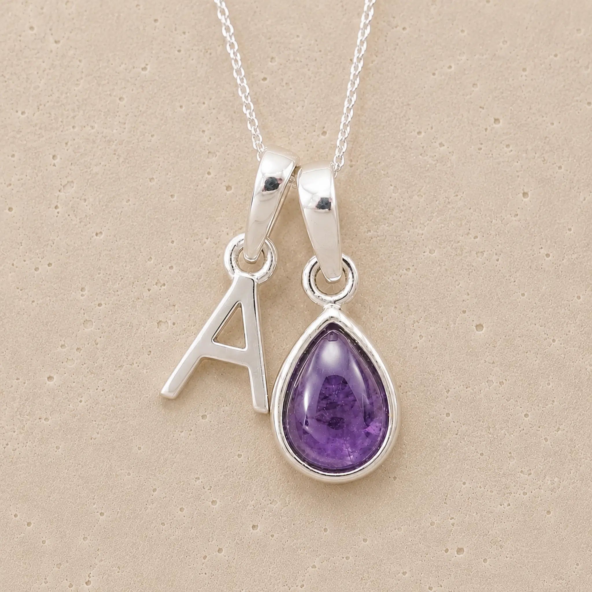 Silver necklace with a teardrop-shaped february birthstone amethyst gemstone pendant and a silver 'A' charm on a beige background.