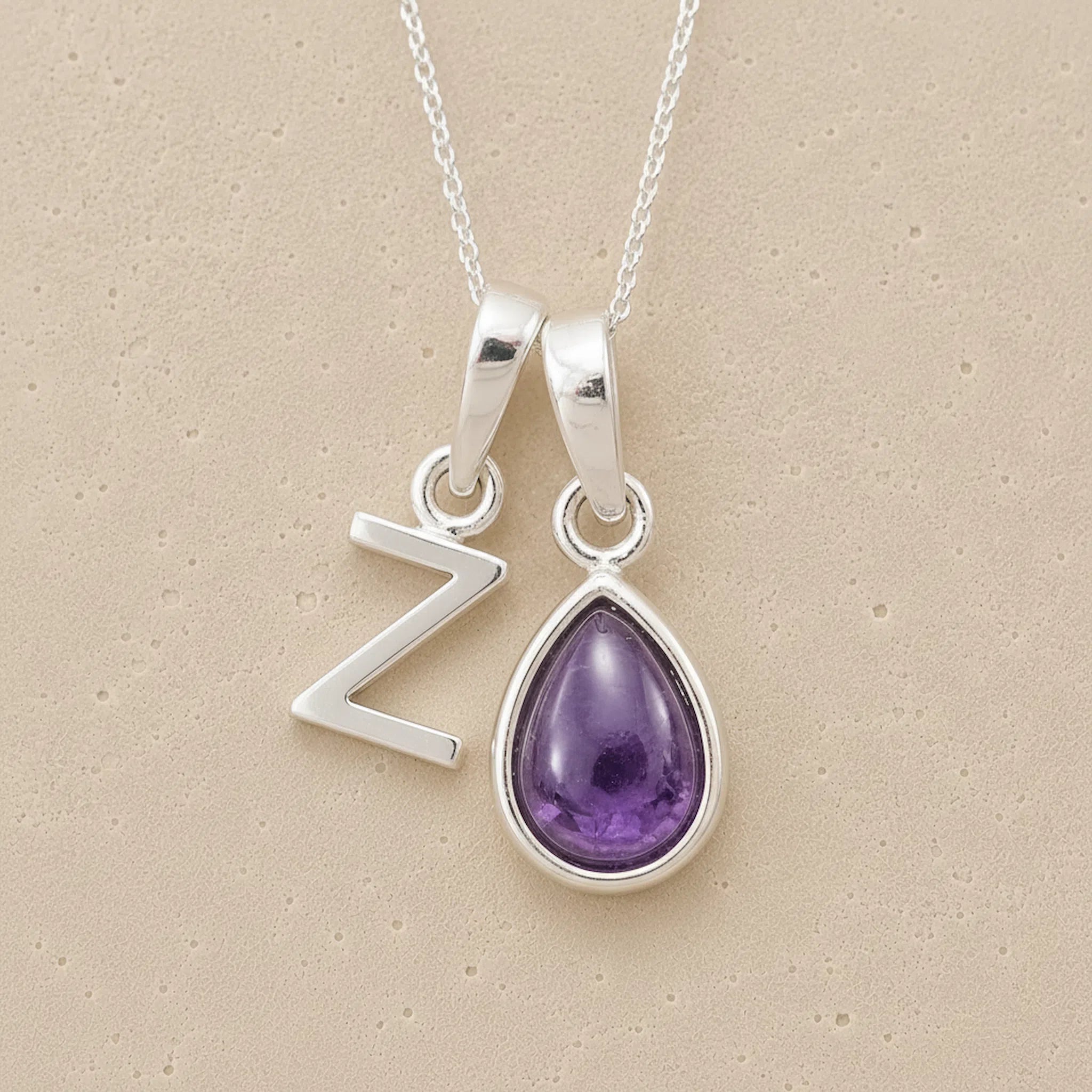 Silver necklace with a teardrop-shaped february birthstone amethyst gemstone pendant and a silver 'Z' charm on a beige background.
